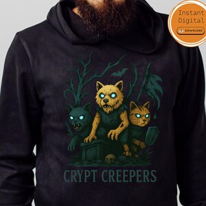 Könnte beinhalten: Schwarzer Kapuzenpullover mit einer Grafik von drei katzenartigen Kreaturen mit leuchtenden Augen und dem Text "CRYPT CREEPERS". Das Design umfasst einen Grabstein, Schädel und einen gruseligen Waldboden. Der Hoodie hat eine Kapuze mit Kordelzug und gerippte Bündchen.