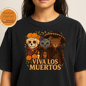 Könnte beinhalten: Schwarzes T-Shirt mit einer farbenfrohen Illustration für den Día de los Muertos. Das Design zeigt einen Hund und zwei Katzen in festlicher Kleidung, mit dem Schriftzug "Viva Los Muertos" in Gold. Das T-Shirt hat einen klassischen Rundhalsausschnitt.