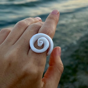 May include: A white, spiral-shaped ring is displayed on a finger against a backdrop of the ocean and sky. The ring's design resembles a seashell or a stylized swirl, offering a unique and artistic aesthetic. The ring is a statement piece.