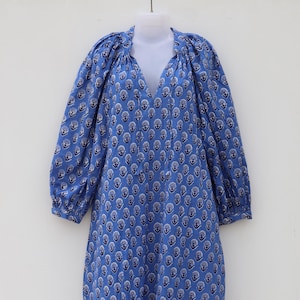 Może przedstawiać: Niebieska sukienka z dekoltem w serek i długimi, pękatymi rękawami. Sukienka jest wzorzysta z powtarzającym się kwiatowym wzorem w kolorze białym i granatowym. Sukienka ma długą, zwiewną sylwetkę.