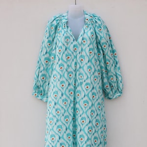 Puede incluir: Un vestido largo y fluido con cuello en V y mangas tres cuartos. El vestido presenta un patrón repetitivo de formas turquesas y blancas con pequeños acentos naranjas. La tela parece ligera y adecuada para climas cálidos.