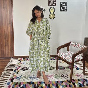Puede incluir: Un vestido largo verde con estampado floral y mangas cortas. El vestido lo lleva una persona de pie sobre una alfombra colorida. A la derecha hay una silla de madera con asiento floral. La pared tiene letreros decorativos que dicen "This is our happy place".