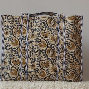 Peut inclure: Un grand sac fourre-tout avec un motif floral dans les tons beige, bleu marine et jaune moutarde. Le sac a deux poignées et est bordé d'un tissu rayé bleu marine et blanc. Le sac est posé sur une surface blanche texturée.