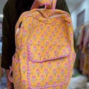 May include: A yellow floral print backpack with pink and white striped trim. The backpack features a front pocket and adjustable straps. The floral pattern is repeated across the bag. The backpack is made of a quilted material.