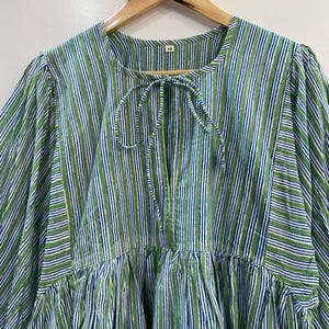 Op de afbeelding: Een groen, wit en blauw gestreepte tuniek top met een strikhalslijn. De top hangt aan een houten hanger. De stof heeft een verticaal streepjespatroon. De top heeft een geplooide taille en lange mouwen.