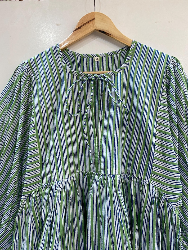 Op de afbeelding: Een groen, wit en blauw gestreepte tuniek top met een strikhalslijn. De top hangt aan een houten hanger. De stof heeft een verticaal streepjespatroon. De top heeft een geplooide taille en lange mouwen.