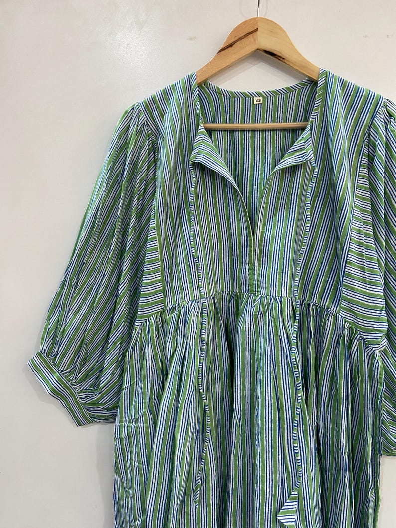 Op de afbeelding: Een groen, wit en blauw gestreepte jurk met driekwart mouwen hangt aan een houten hanger. De jurk heeft een geplooid taille en een split halslijn met een striksluiting. De stof heeft verticale strepen.