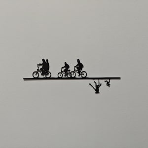 Op de afbeelding: Zwart metalen wandkunst met silhouetten van mensen op fietsen en figuren die eronder vallen. Het kunstwerk heeft een minimalistisch ontwerp en is geschikt voor interieurdecoratie.