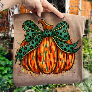 Peut inclure: Un coussin marron avec un motif de citrouille colorée. La citrouille est orange avec des accents dorés, verts et rouges, et un nœud vert à pois noirs. Le coussin est tenu par une main.