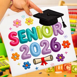 Peut inclure: Tissu blanc avec texte et graphiques brodés. Le mot "SENIOR" est en lettres colorées, avec "2026" en dessous. Une toque noire et un parchemin sont également brodés. Des motifs floraux et des étoiles complètent le design.