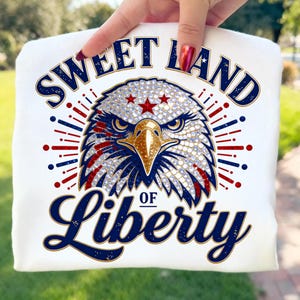 Peut inclure: Sweat-shirt blanc avec un motif détaillé de tête d'aigle. L'aigle présente des ornements scintillants, des accents rouges et bleus et le texte bleu marine "SWEET LAND OF LIBERTY". Un design patriotique.