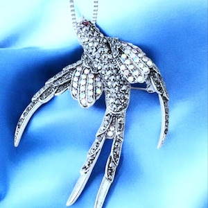May include: A silver-toned bird pendant necklace with a detailed design. The bird is encrusted with clear and iridescent crystals, with a small red stone for the eye. The wings are spread in flight, and the pendant hangs from a delicate chain.