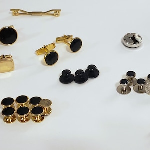 May include: A collection of gold-toned and silver-toned men's accessories, including cufflinks, tie bars, and shirt studs. The cufflinks feature black circular and square designs. The tie bar is a simple, elegant design. The shirt studs have black and silver accents.