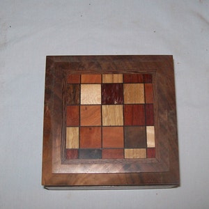 May include: A wooden box with a decorative inlay of various wood types in a checkered pattern. The box is made of dark wood and has a light brown border.