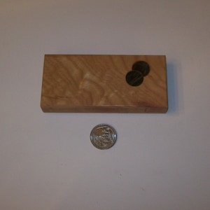 Könnte beinhalten: Eine kleine Holzkiste mit einer hellbraunen Holzmaserung. Die Kiste hat eine kleine Vertiefung oben, in der zwei Münzen liegen.