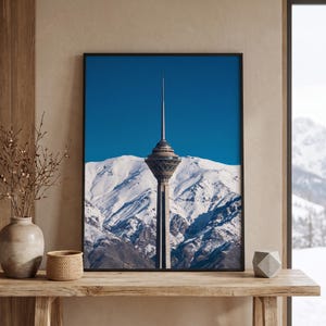 May include: A framed wall art print featuring a tall, slender communication tower with a wide, circular observation deck at the top, set against a backdrop of snow-covered mountain peaks under a clear, deep blue sky during the daytime.
