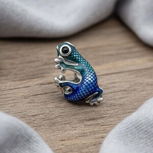 Può includere: Un anello a forma di rana in argento con finitura smaltata blu e verde. La rana ha grandi occhi neri ed è posizionata come se fosse seduta. L'anello è su una superficie di legno con un panno grigio sullo sfondo.