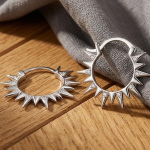 May include: A pair of silver-toned hoop earrings featuring a sunburst design with pointed spikes radiating outwards. Each spike is accented with a small, clear, sparkling stone embedded near the base, creating a bright, reflective metallic finish.