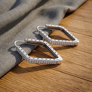 May include: A pair of silver-toned hoop earrings in a diamond shape. The front surface of each earring is encrusted with small, clear, sparkling stones, while the outer edges feature a repeating pattern of tiny, engraved heart shapes.