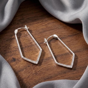 Puede incluir: Un par de pendientes de aro geométricos plateados. Los pendientes tienen forma rectangular con la parte inferior puntiaguda y están incrustados con pequeñas piedras brillantes a lo largo del borde inferior. Los pendientes se muestran sobre una superficie de madera.