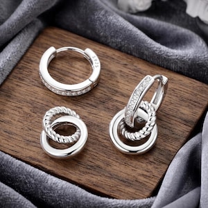 May include: Three pairs of silver earrings on a wooden surface. One pair is a classic hoop design with small, clear stones. Another features two intertwined hoops, and the third combines a hoop with a braided design.
