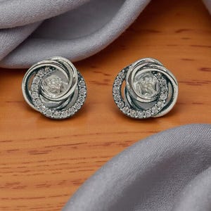 May include: A pair of silver stud earrings. Each earring has a swirling design with a circle of small, clear gemstones around the edge. The earrings are on a wooden surface with grey fabric.