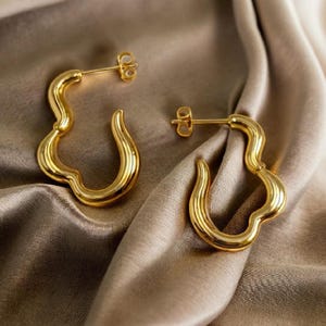 May include: A pair of gold-colored hoop earrings with a unique, wavy design. The earrings have a post closure and a smooth, polished finish. The earrings are displayed on a satin fabric.