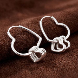Puede incluir: Un par de pendientes de aro de plata con forma de corazón. Cada pendiente presenta un aro grande en forma de corazón con tres pequeños dijes colgantes también en forma de corazón en la parte inferior. El metal tiene grabado el texto S925.