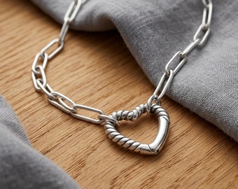 May include: A silver-toned chain necklace featuring elongated rectangular links and a central heart-shaped pendant. The pendant has a twisted rope-like texture and functions as a clasp, connecting the two ends of the chain.