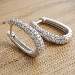 May include: A pair of silver-toned hoop earrings with an elongated oval shape. The front-facing surface of each hoop is encrusted with multiple small, clear, sparkling stones arranged in a dense, multi-row pavé setting, creating a bright, reflective finish.