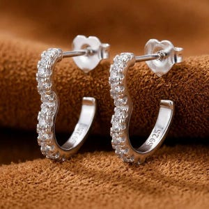 May include: A pair of silver hoop earrings in a heart shape, featuring a row of small clear stones along the front. The earrings have a post back closure and are stamped with S925 on the inner band, indicating sterling silver construction.