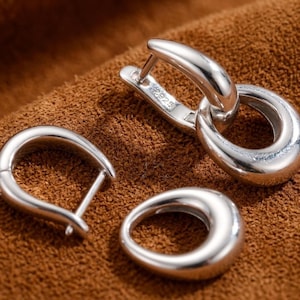 Pode incluir: Um par de brincos de argola em prata polida com um formato de gota suave e arredondado. Um brinco é exibido com o fecho articulado aberto, revelando uma pequena marca 925 na superfície interna, enquanto o outro brinco está fechado.