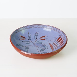 May include: A handmade ceramic bowl with a reddish-brown exterior and a light blue interior. The interior features a floral design in blue and red, with the words "MERCY SHALL FOLLOW" around the rim. A decorative piece for the home.