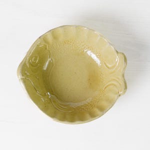 May include: A ceramic dish in the shape of a fish, with a light yellow-green glaze. The dish has a scalloped edge and embossed details, including a fish eye and fin patterns. The dish is on a white surface.