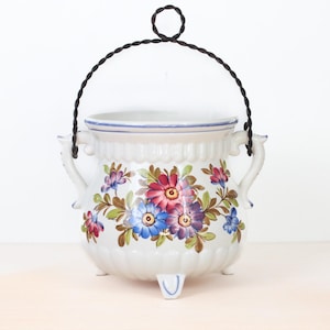 Puede incluir: Jardinera colgante de cerámica blanca con asa de metal negro. La jardinera está decorada con diseños florales coloridos en rojo, azul y morado. Tiene tres patas pequeñas y dos asas.