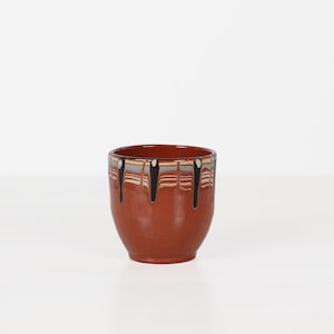 May include: A terracotta-colored ceramic cup featuring a smooth, rounded body. The upper rim is decorated with a horizontal band of grey and white stripes, accented by vertical black drip-like glaze patterns that extend downward from the top edge.