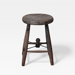 May include: A rustic, three-legged wooden stool with a round seat. The stool is made of weathered wood, showing signs of age and use. The legs are slightly splayed, and a crossbar provides stability. The stool's height is approximately 18 inches.
