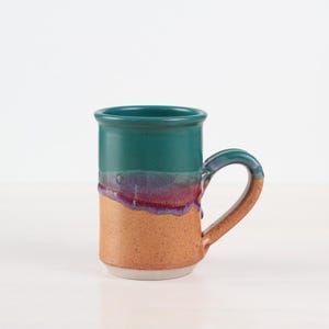 May include: A handmade ceramic mug with a teal rim and handle, transitioning to purple and red hues, and a tan base. The mug has a cylindrical shape and a curved handle. This unique mug is perfect for coffee or tea.