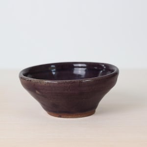 May include: A small, handmade ceramic bowl with a deep purple glaze. The bowl has a slightly irregular shape and a textured surface. The interior is smooth and glossy, while the exterior has a more rustic appearance. The bowl is suitable for serving small portions of food.