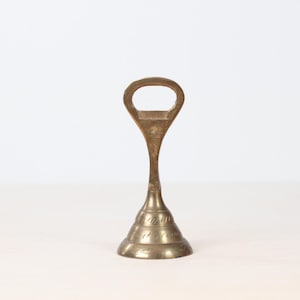 May include: A vintage brass bottle opener with a distinctive design. The opener features a loop handle and a conical base with etched detailing. The brass has a warm, aged patina, suggesting its vintage origin. The opener stands upright against a neutral background.