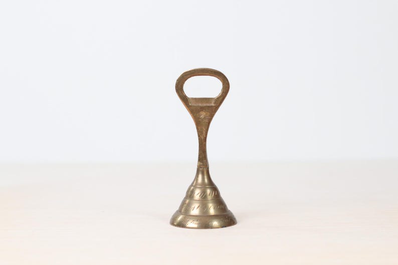 May include: A vintage brass bottle opener with a distinctive design. The opener features a loop handle and a conical base with etched detailing. The brass has a warm, aged patina, suggesting its vintage origin. The opener stands upright against a neutral background.