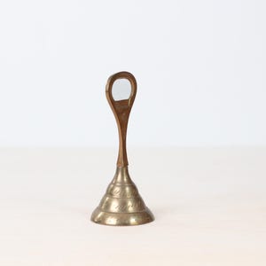 May include: A vintage brass bottle opener with a teardrop-shaped handle and a conical base. The base features etched decorative patterns. The opener stands upright on a light-colored surface against a white background.