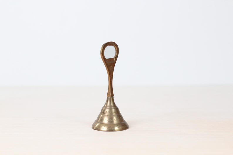 May include: A vintage brass bottle opener with a teardrop-shaped handle and a conical base. The base features etched decorative patterns. The opener stands upright on a light-colored surface against a white background.