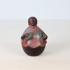 Puede incluir: Figura de piedra tallada a mano de una persona sosteniendo un objeto verde. La figura tiene una base marrón oscuro y un cuerpo con tonos rojos, verdes y marrones. La cara tiene una expresión sonriente.