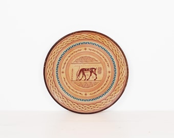 May include: A round ceramic plate featuring a central illustration of a four-legged animal in dark red, surrounded by concentric rings of geometric patterns, a blue and white Greek key border, and an outer rim with a tan braided rope design.