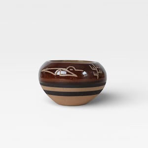 May include: A small, round ceramic bowl with a glossy brown finish. The bowl features a tan and black striped design around the base and a carved bird and symbol design near the top. The bowl is approximately 4 inches in diameter.