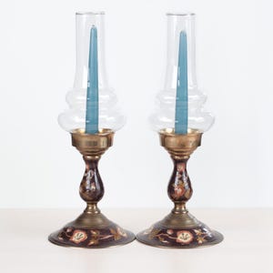 May include: Two antique candle holders with clear glass hurricane shades. Each holder has a teal candle inside. The bases are dark brown with floral accents and brass details. The candle holders are on a white surface.