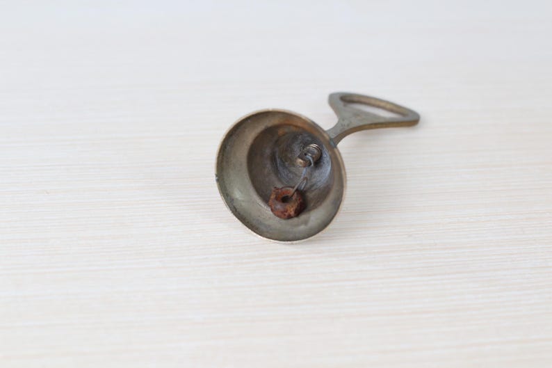 May include: A vintage brass bell with a handle, resting on a light-colored surface. The bell has a rounded shape with a small clapper inside. The handle is arched, and the bell appears aged.