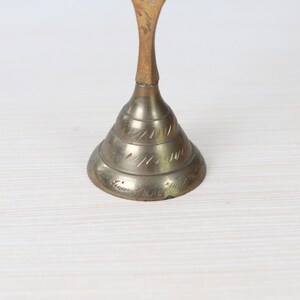 May include: A vintage brass bell with a conical base and a slender, curved handle. The bell's base features etched decorative lines. The handle is a lighter color than the bell. The bell is likely used for decorative purposes.