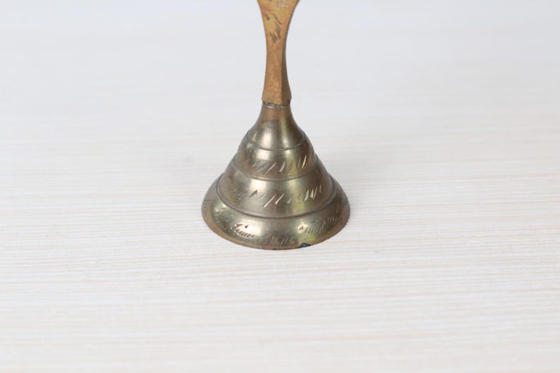 May include: A vintage brass bell with a conical base and a slender, curved handle. The bell's base features etched decorative lines. The handle is a lighter color than the bell. The bell is likely used for decorative purposes.